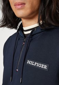 Felpa navy con design a mezza zip, caratterizzata da un patch logo testurizzato, dettagli in coste sul collo e cappuccio con lacci e punte in metallo.