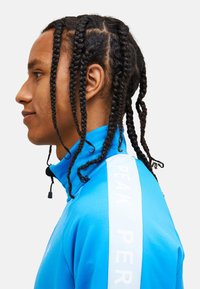 Giacca sportiva blu brillante con colletto alto, dettaglio con cordino e branding bianco "PEAK PERFORMANCE" sulla spalla. Tessuto strutturato.