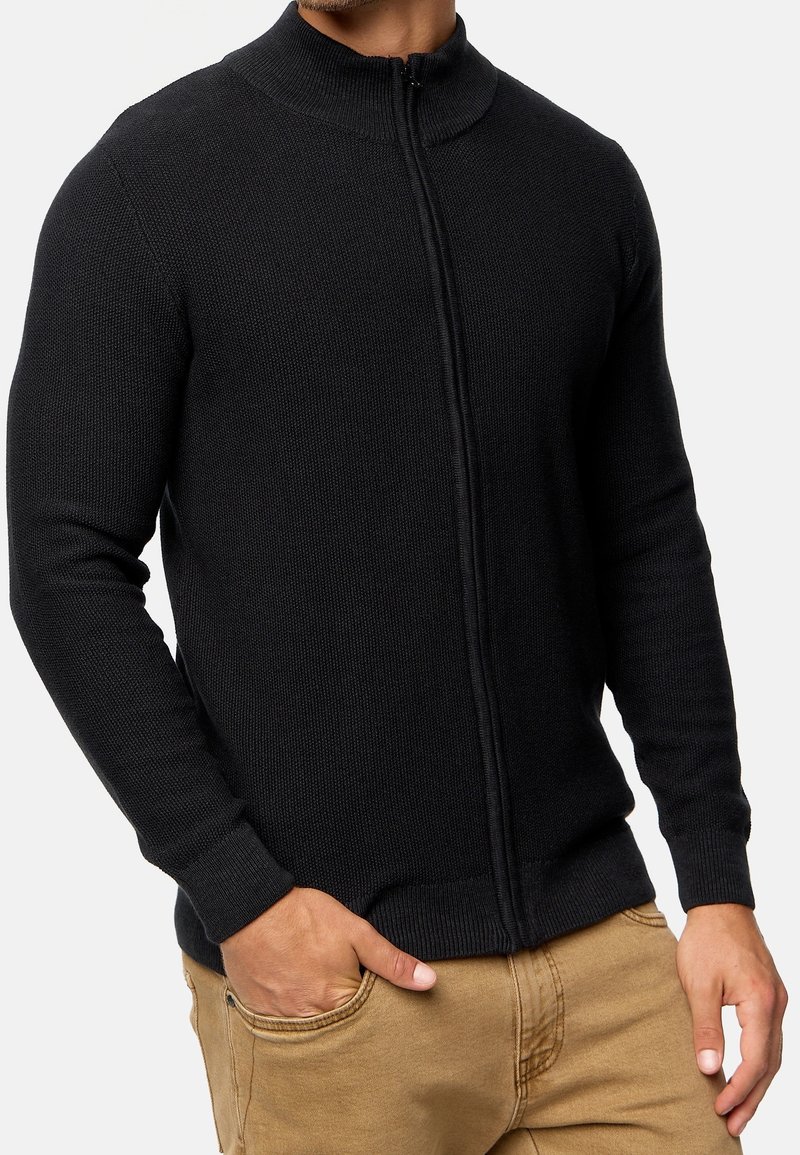 Mann trägt einen schwarzen, vollständig zugeknöpften Strickpullover mit den Händen in den Taschen einer beigen Hose, zeigt Oberkörper und Torso vor einem weißen Hintergrund.