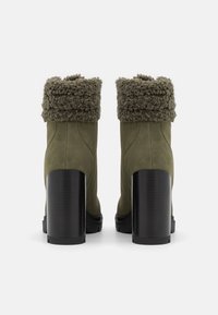 Para oliwkowych zamszowych botków za kostkę z grubymi czarnymi blokowymi obcasami i złożonymi brązowymi mankietami z sztucznego futra, pokazanych z tyłu.