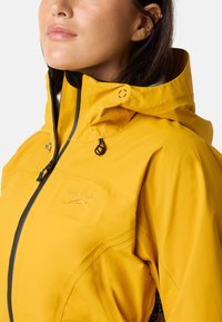 Gul vandtæt jakke med lynlås foran, justerbar hætte og sorte detaljer. Har logo-broderi og tekstureret stof.
