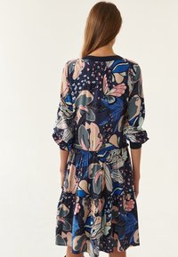 Kleid mit Blumenmuster, das einen marineblauen Grundton, lange Ärmel, eine geraffte Taille und einen fließenden, gefransten Saum mit rosafarbenen und grünen Akzenten aufweist.