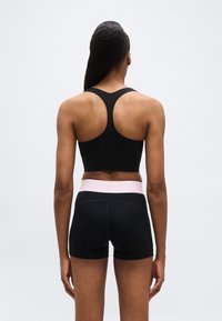 Femme debout, de dos, portant un soutien-gorge de sport noir à dos nageur et un short noir avec une ceinture rose sur un fond uni