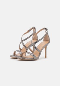 Silver glittrande högklackade sandaler med crisscross-remdesign, öppen tå och ankelspänne. Slät innersula med stilettklack.