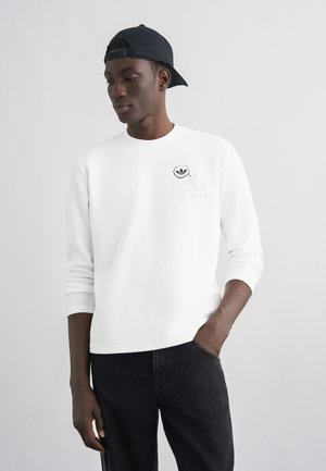 Giovane uomo con una maglia bianca a maniche lunghe con un piccolo logo e un berretto da baseball nero indossato al contrario, in piedi con una mano in tasca.