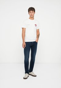 Camiseta blanca de algodón con un diseño gráfico rojo y negro en el lado izquierdo del pecho, combinada con unos vaqueros de mezclilla azul oscuro y zapatillas de color claro.