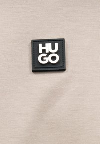 Μαύρο τετράγωνο μπαλώμα με ανάγλυφα λευκά γράμματα "HU GO" σε ανοιχτό μπεζ ύφασμα.