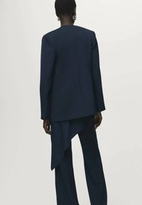 Laivastonsininen räätälöity puku, joka sisältää pitkän, epäsymmetrisen bleiserin ja yhdistelevät leveät housut, valmistettu sileästä kankaasta, jolla on jämäkkä siluetti.