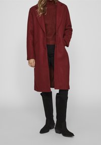 Long manteau en laine rouge avec revers et poches latérales, associé à un pull rouge texturé et un pantalon noir ajusté, complété par des bottes noires montant jusqu'aux genoux.