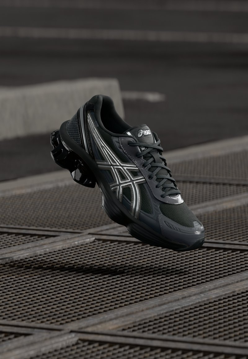 Chaussure de course Asics noire et grise avec une semelle épaisse et des lacets, positionnée en l’air au-dessus d’une surface en grille métallique dans un environnement urbain.