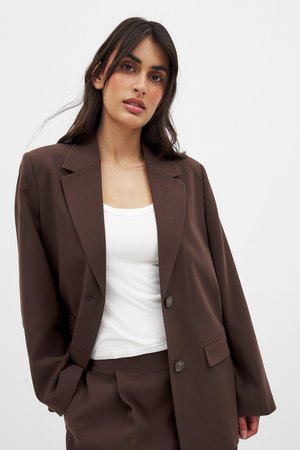 Brun oversized blazer med slag, to knapper og sidelommer, båret over en hvid tanktop. Glat stof med en skræddersyet pasform.
