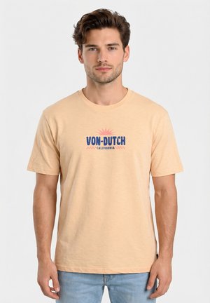 Giovane uomo con capelli corti e ondulati che indossa una T-shirt beige con la scritta "VON-DUTCH CALIFORNIA" e jeans azzurri, in piedi davanti a uno sfondo semplice.