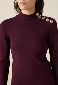 Femme portant un pull en maille côtelée, de couleur violet foncé, à manches longues avec un col montant et cinq boutons dorés sur l'épaule gauche.