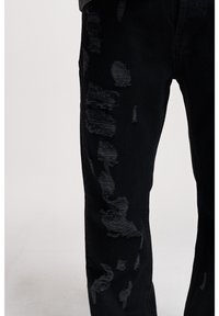 Jean noir en denim avec des détails usés et des patches effilochés de différentes tailles. Le matériau est texturé avec un léger brillant.