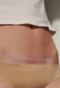 Beige ondergoed met een gladde, transparante kleefpatch die horizontaal op de onderbuik is geplaatst. Huidskleur is zichtbaar rondom de patch.