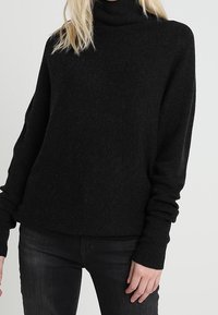 Pull à col roulé noir en tissu tricoté texturé, doté d'une coupe décontractée et de manches longues, associé à un jean foncé.