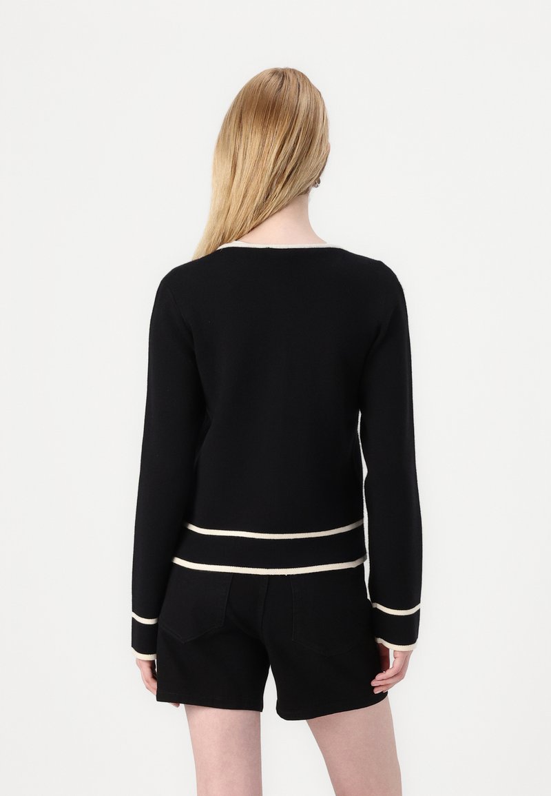 Maglione nero con maniche lunghe, strisce orizzontali bianche in vita e sui polsini, collo rotondo e texture liscia. Indossato con pantaloni corti neri.