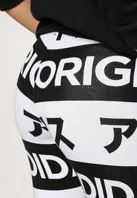 Černobílé legíny s velkými textovými vzory v tučném písmu. Materiál vypadá elasticky a má hladkou texturu.