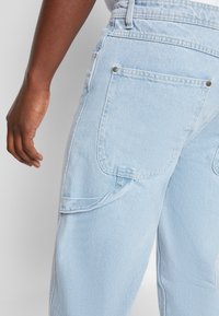 Light blue denim pants with a high waist, featuring two back pockets and reinforced stitching along the seams. Smooth texture, casual design.
