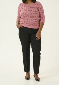 Rosa gestrickter Pullover mit gemustertem Textur und gerippten Bündchen, kombiniert mit schwarzen, maßgeschneiderten Hosen. Das Ensemble umfasst schwarze spitze Schuhe.