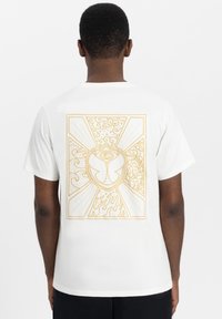 Witte katoenen t-shirt met een gouden geometrisch ontwerp met een oog en gestileerde patronen op de achterkant, korte mouwen en een ronde halslijn.