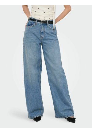 Personne portant un jean bleu clair à jambes larges avec une ceinture noire et des chaussures noires pointues, debout les mains dans les poches.