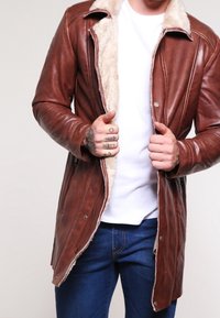 Uomo che indossa una giacca di montone marrone in pelle sopra una t-shirt bianca e jeans blu, mentre sistema la giacca con le mani tatuate in vista.