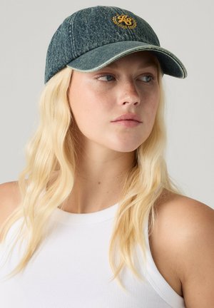 Lyshåret kvinde iført en blå denim kasket med gul broderi og en hvid ærmeløs top, der kigger en smule mod højre.