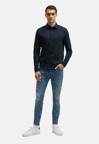Marineblaues Langarmhemd mit Knopfverschluss, aus glattem Stoff. Kombiniert mit hellblauen Jeans und weißen Sneakers. Schlichtes Design und Passform.