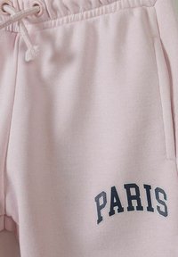 Roza trenirke s pasom na vrvico in stranskimi žepi. Na levi strani je temno modra grafika "PARIS". Mehka, teksturirana tkanina.