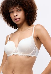 Sujetador beige con encaje, copas acolchadas, tirantes ajustables y bordes festoneados. La tela suave combinada con detalles de encaje floral proporciona un contraste.