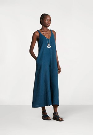 Femme debout portant une combinaison bleue sans manches, des sandales noires à fines lanières et un long collier pendentif sur un fond blanc uni.
