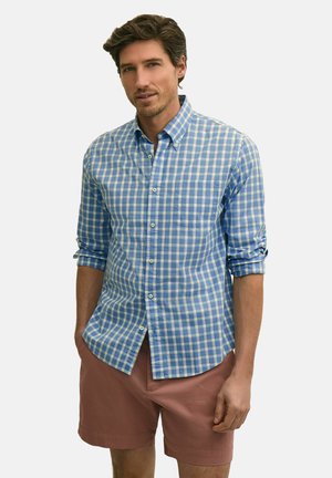 Mann trägt ein blau-weiß kariertes Hemd mit aufgerollten Ärmeln und pinke Shorts, steht vor einem einfarbigen Hintergrund.