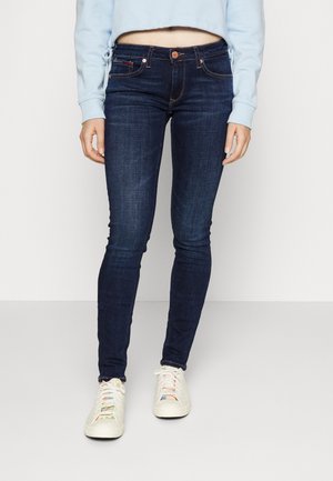 Jeans ajustados de color azul oscuro con un acabado suave, que cuentan con una cinturilla estándar y un cierre de botón, combinados con zapatillas de color claro.