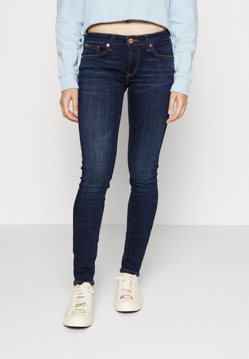Jeans ajustados de color azul oscuro con un acabado suave, que cuentan con una cinturilla estándar y un cierre de botón, combinados con zapatillas de color claro.