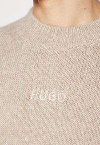 Pull en maille beige avec col côtelé et nom de marque brodé en blanc "HUGO" sur la poitrine, porté par une personne.