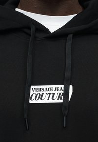 Sweat à capuche noir avec des cordons épais noirs et une étiquette blanche portant l'inscription "VERSACE JEANS COUTURE" sur la poitrine, porté par-dessus une chemise blanche.