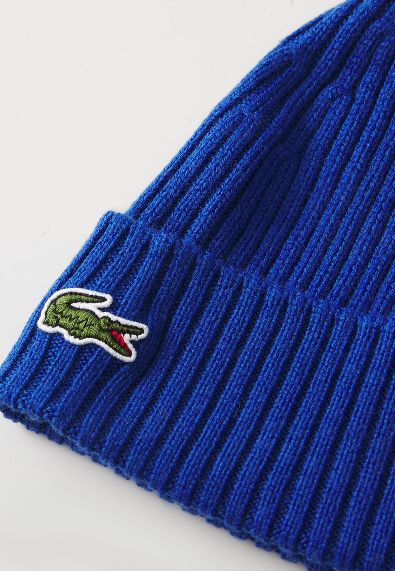 Blauer Strickbeanie mit geripptem Muster und umgeschlagenem Saum. Auf der Seite befindet sich ein grünes Krokodil-Logo, umrahmt von Weiß.
