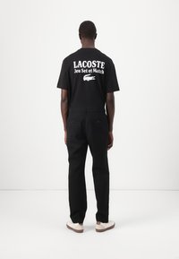 Black Lacoste t-shirt with white text and logo on the back, paired with black pants and white shoes. Simple design, standard fit.