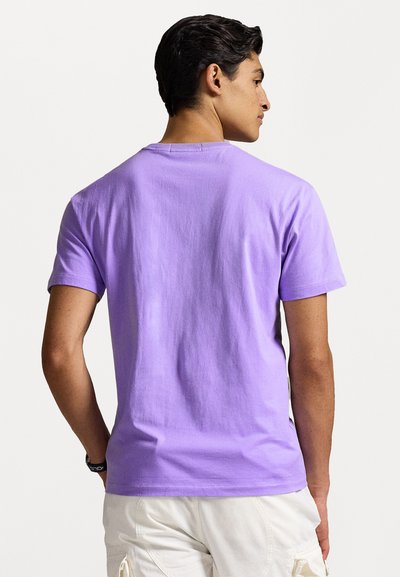 Camiseta de algodón morada con mangas cortas, cuello redondo y una textura suave, mostrada desde la parte posterior, combinada con pantalones cortos de color claro.