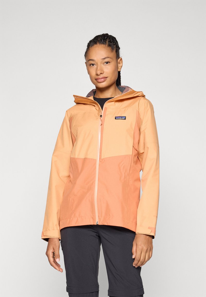 Vattentät jacka med en orange gradient, utrustad med dragkedja fram, huva och en logotyp på bröstet. Slät textur, långa ärmar.