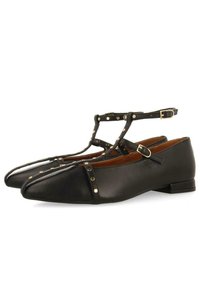 Bailarinas de cuero negro con punta afilada, diseño de tira T, tachuelas en tono dorado y hebilla en el tobillo. Textura suave y tacón bajo tipo bloque.