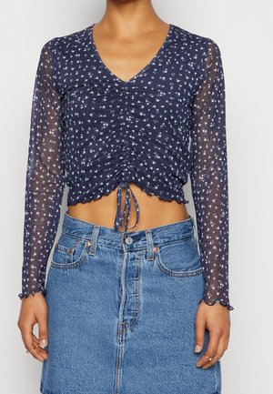 Frau trägt ein marineblaues, florales, transparentes Crop-Top mit langen Ärmeln und einem Kordelzug vorne, kombiniert mit einem hoch taillierten blauen Jeansrock.