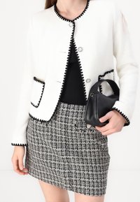 Kvinna i vit strukturerad jacka med svarta detaljer, svart topp, svart och vit rutad kjol, som håller en liten svart läderhandväska.