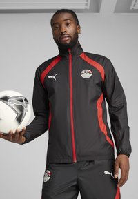 Veste de sport noire avec des accents rouges, dotée d'une fermeture éclair, d'un logo et d'un emblème, associée à un pantalon noir et tenant un ballon de football dans la main.