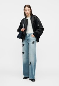 Γυναίκα με μαύρο bomber μπουφάν, λευκό ριμπ μπλουζάκι, ανοιχτό μπλε τζιν παντελόνι με φαρδύ μπατζάκι και μαύρα μοτίβα αστέρια, και μαύρα παπούτσια, κρατώντας μια μαύρη τσάντα χειρός.