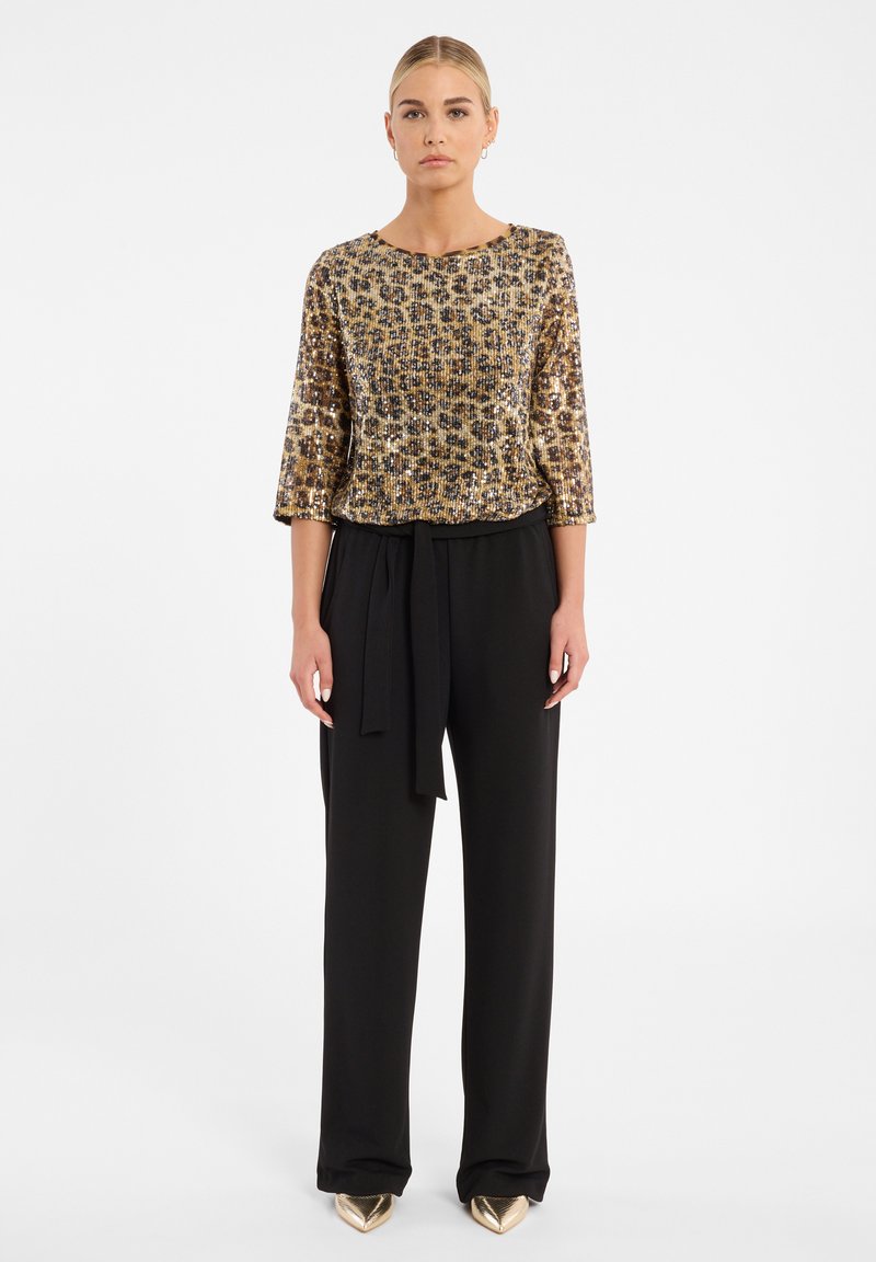 Haut à sequins dorés et noirs avec des manches trois-quarts, associé à un pantalon noir taille haute avec une ceinture nouée. Chaussures dorées à bout pointu.