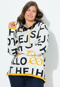 Femme aux cheveux bruns ondulés portant un sweatshirt blanc avec des lettres noires en gras et des accents jaunes, souriante et tenant un cordon de sweatshirt.