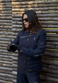 Vrouw met zonnebril, marineblauwe gewatteerde jas met bruine zoom en zwarte leren handschoenen, staand bij een houten omheining in daglicht.