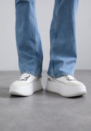 White sneakers with a platform sole, textured detailing, and black accents, paired with light blue straight-leg jeans.